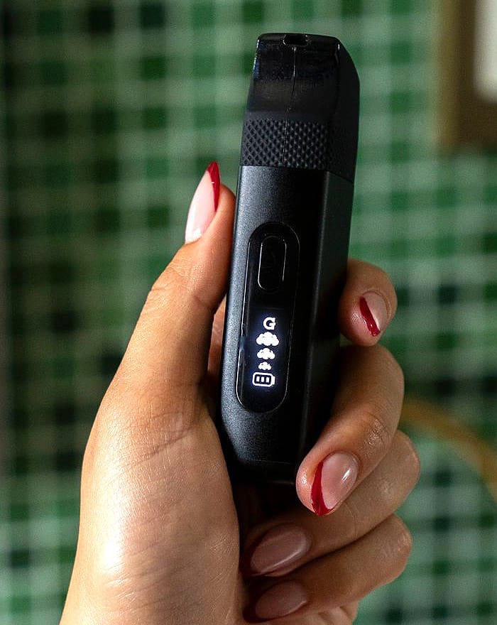 Hand holding the G Pen Micro II showing its LED screen on a green background